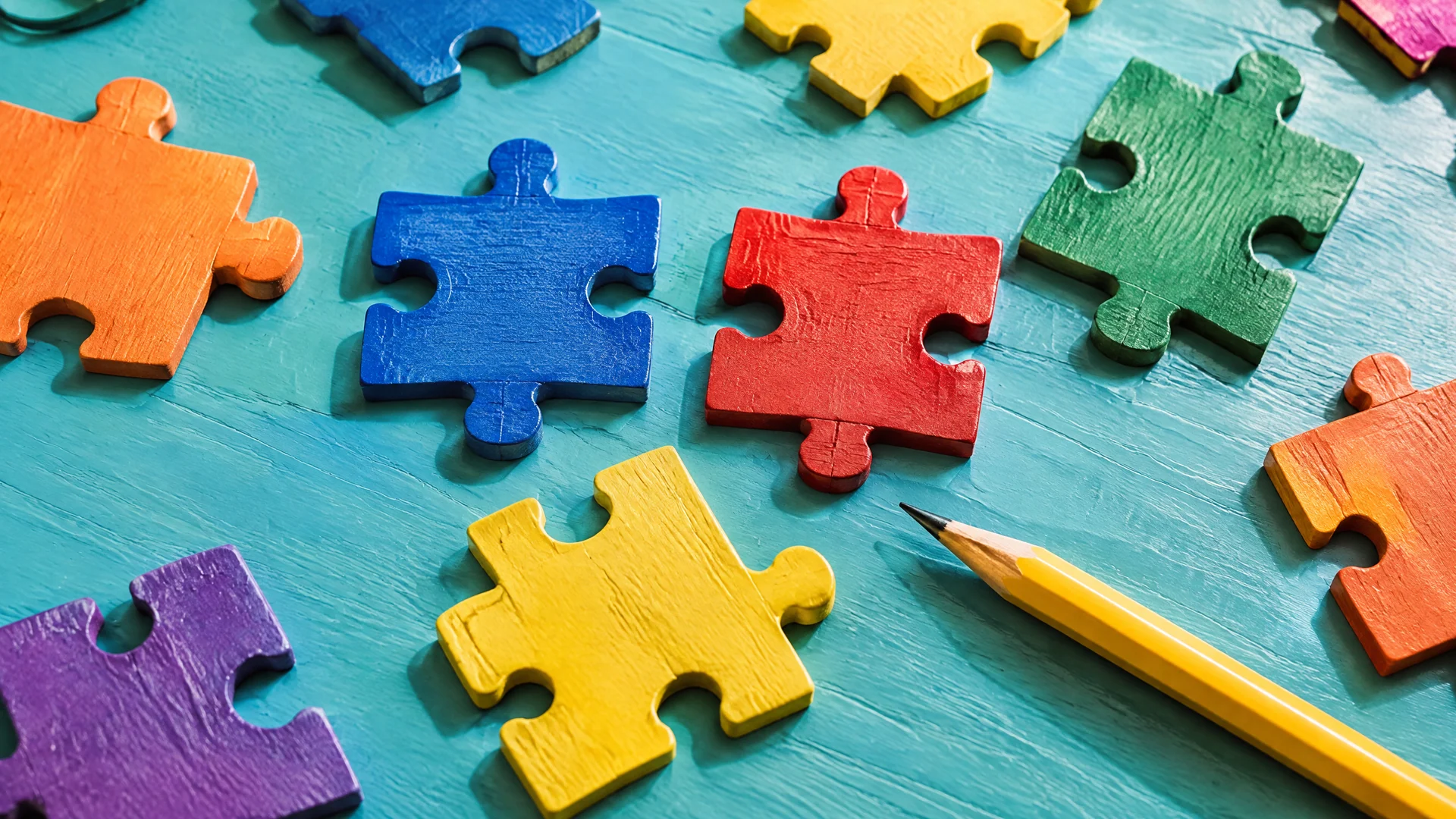 Brightly painted wooden puzzle pieces and a yellow pencil on a teal table